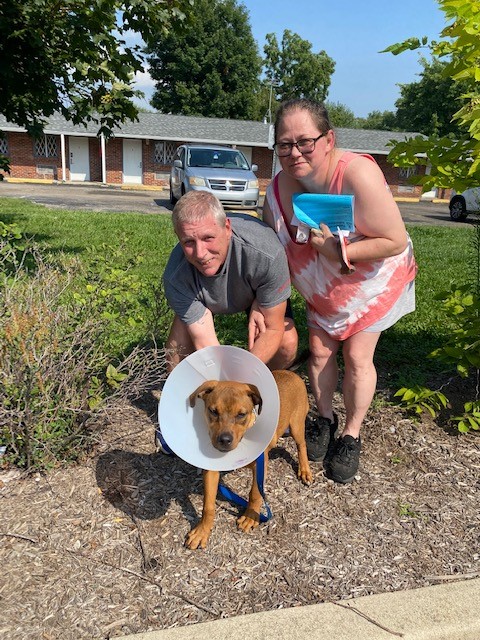 Man and woman posing with a brown dog wearing a cone collar, outside a building