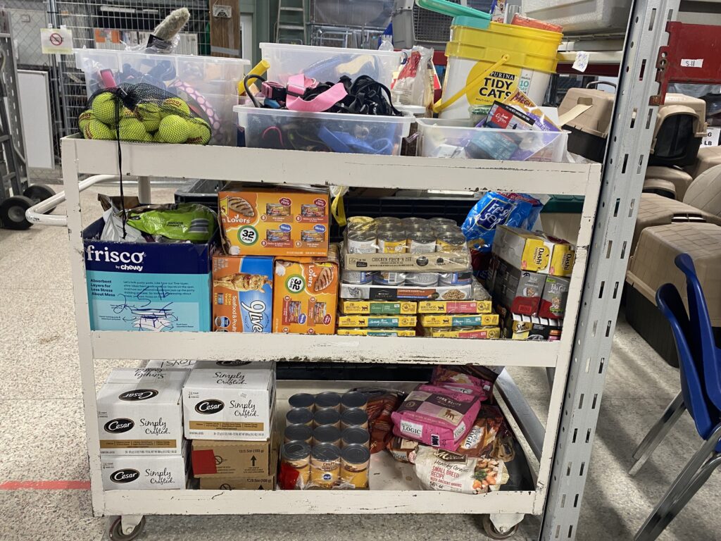 A cart filled with various pet supplies, including tennis balls, cat food, dog food, and cleaning supplies in a storage area