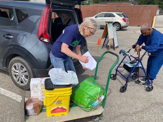 Volunteer unloading pet supplies from a car onto a cart while a woman with a walker waits