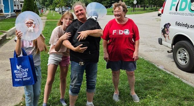 A family with a dog wearing a cone collar is standing by a FIDO Indy van.