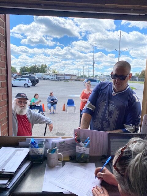 A man in a Dallas Cowboys jersey interacting with a volunteer at the FIDO Indy facility, with other people waiting outside