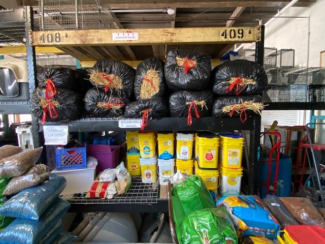 Shelves stacked with large bags of hay wrapped in black plastic and multiple containers of cat litter in a storage area